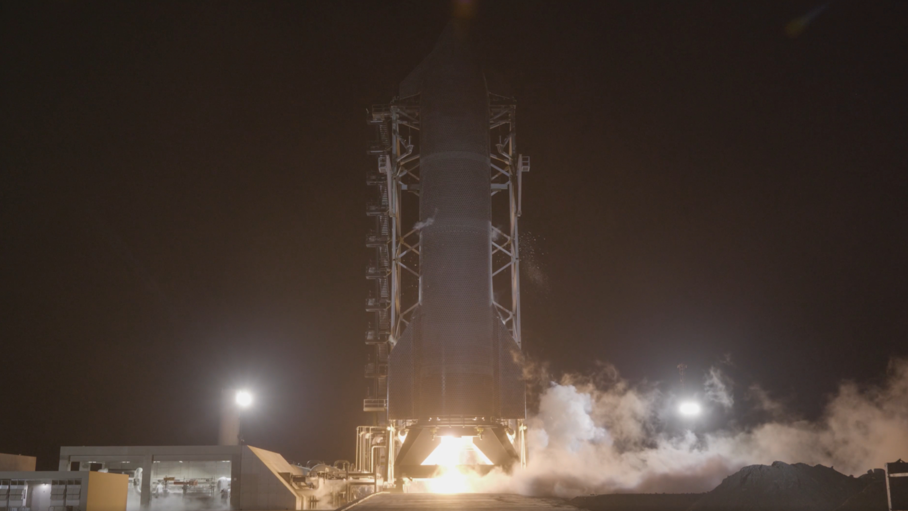 A tall finned black rocket fires an engine on a test stand in the black of night.