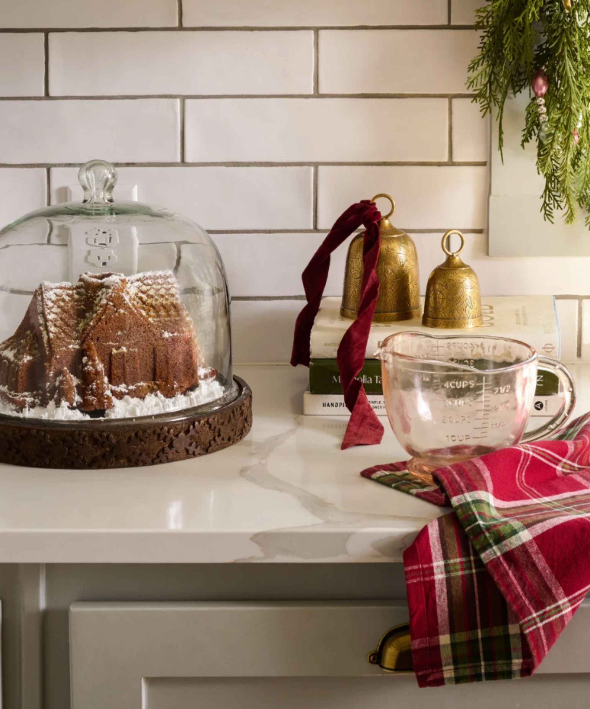 White marble counter with red plaid dish towel, blush glass measuring jug, brass bell ornaments and glass dome with gingerbread house inside