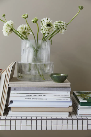 clear ribbed glass vase with white flowers on top of a stack of magazines