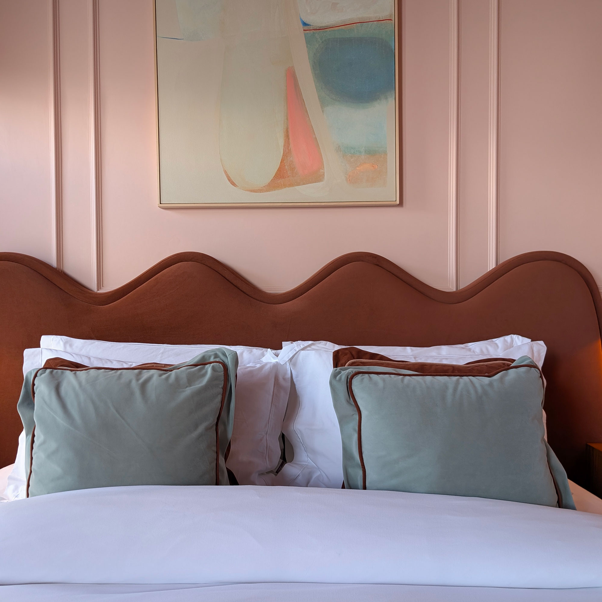 Close up of brown wavy bed headboard in front of pink panelled wall with pastel artwork