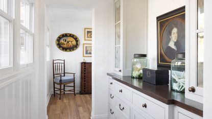 Galley kitchen with white cabinetry, black countertops, glass-fronted cabinets, an antique portrait, glass jars filled with shells, and a vintage wooden chair with vintage art above it.