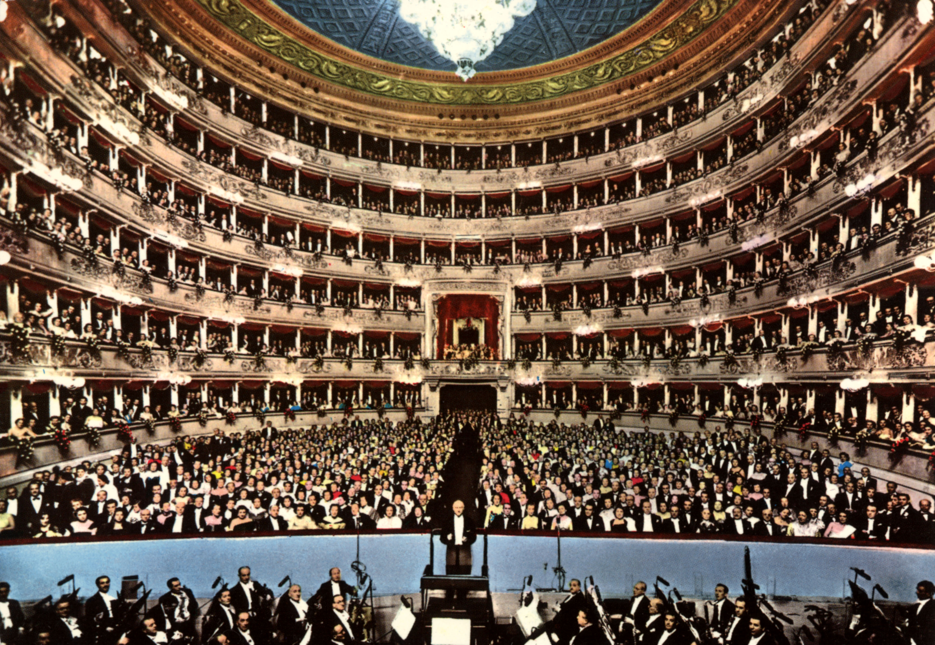 A packed opera house viewed from the stage, with hundreds of elegantly dressed audience members filling the stalls and tiered balconies beneath an ornate domed ceiling.
