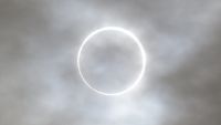 The edge of the sun is visible around the silhouette of the moon during an eclipse, forming a burning ring behind a veil of thin clouds.