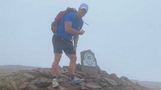 A man stands at the top of Pen y Fan, pointing at the National Trust sign, in the middle of completing the Fan Dance.