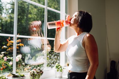 Woman drinking water wearing activewear, standing by window with sunshine coming in