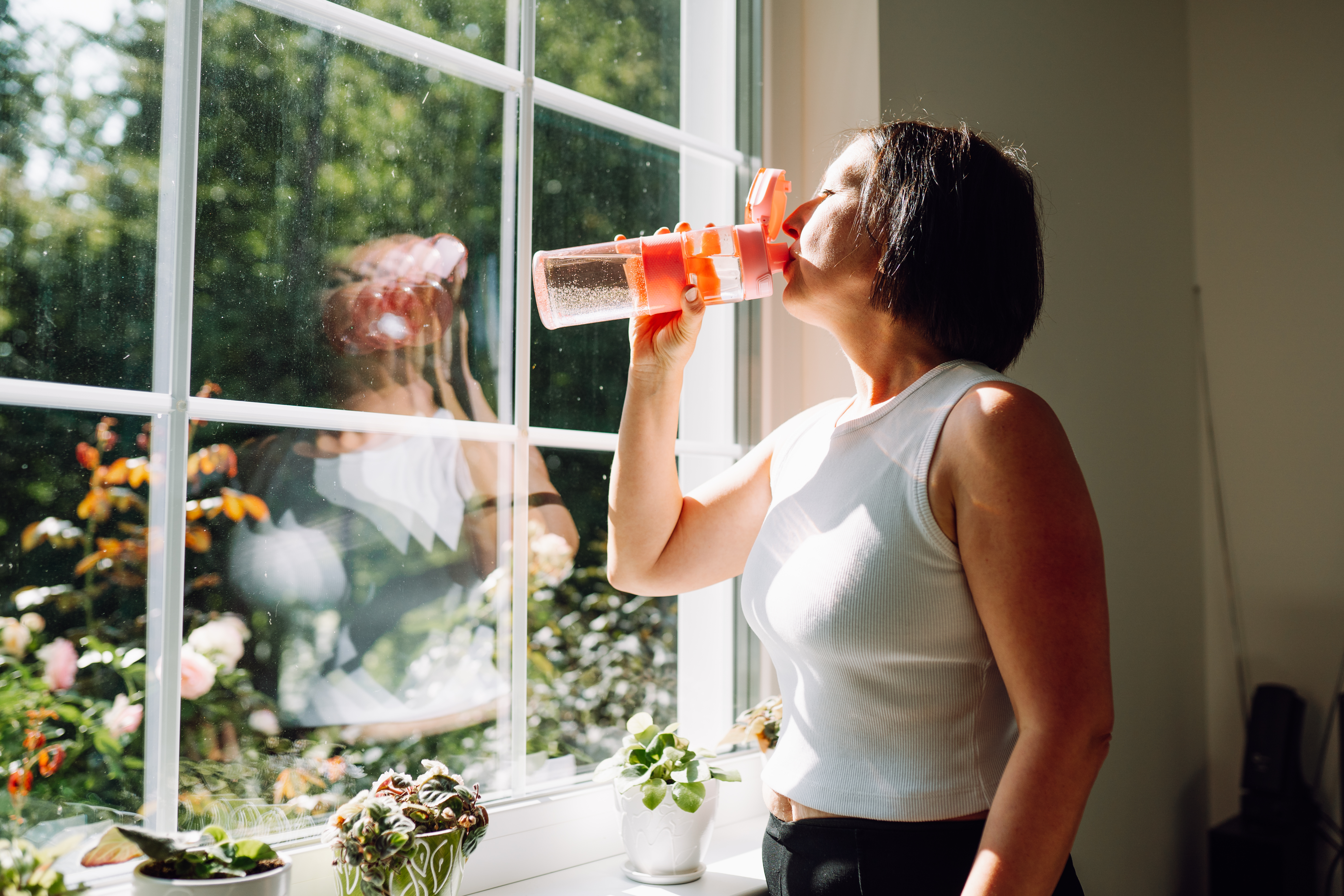 Woman drinking water wearing activewear, standing by window with sunshine coming in