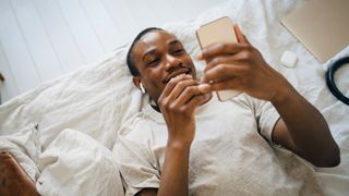 A smiling man scrolls through phone looking for sleep podcasts while lying in bed