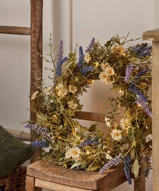A green floral wreath with green stems, lavender, and daisies decorated on it, and a wooden rustic chair with a rustic wooden ladder and basket with a fuzzy dark green pillow to the left of it