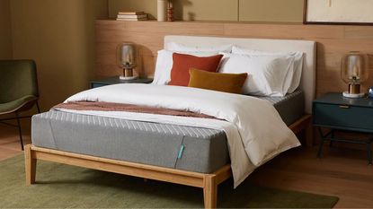 A grey Siena mattress pictured on a white bed. with a wooden base, against a wooden panelled wall. The flooring is the same light wood with a green rug, and to either side of the bed are matching blue bedside tables, with lamps. To the left, there's also a green chair. Above the wood panelling, the wall is pale green.