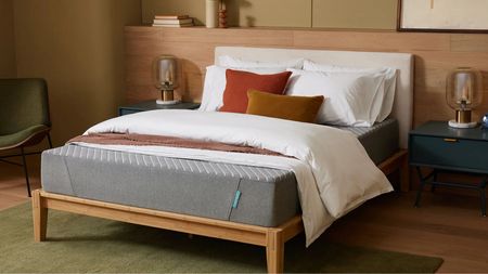 A grey Siena mattress pictured on a white bed. with a wooden base, against a wooden panelled wall. The flooring is the same light wood with a green rug, and to either side of the bed are matching blue bedside tables, with lamps. To the left, there's also a green chair. Above the wood panelling, the wall is pale green.