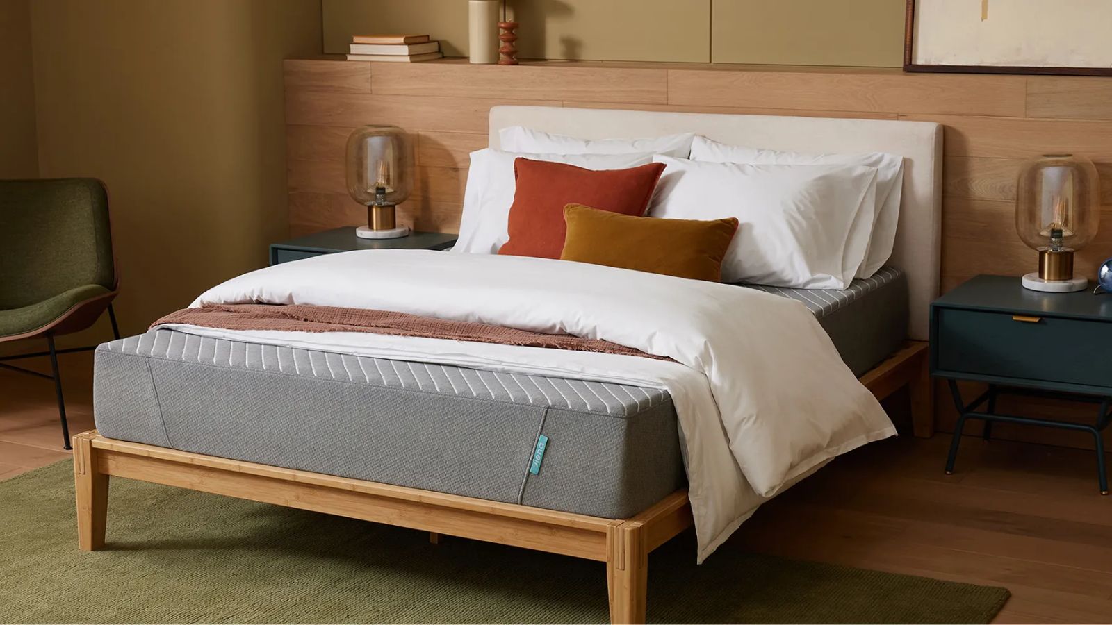 A grey Siena mattress pictured on a white bed. with a wooden base, against a wooden panelled wall. The flooring is the same light wood with a green rug, and to either side of the bed are matching blue bedside tables, with lamps. To the left, there's also a green chair. Above the wood panelling, the wall is pale green.