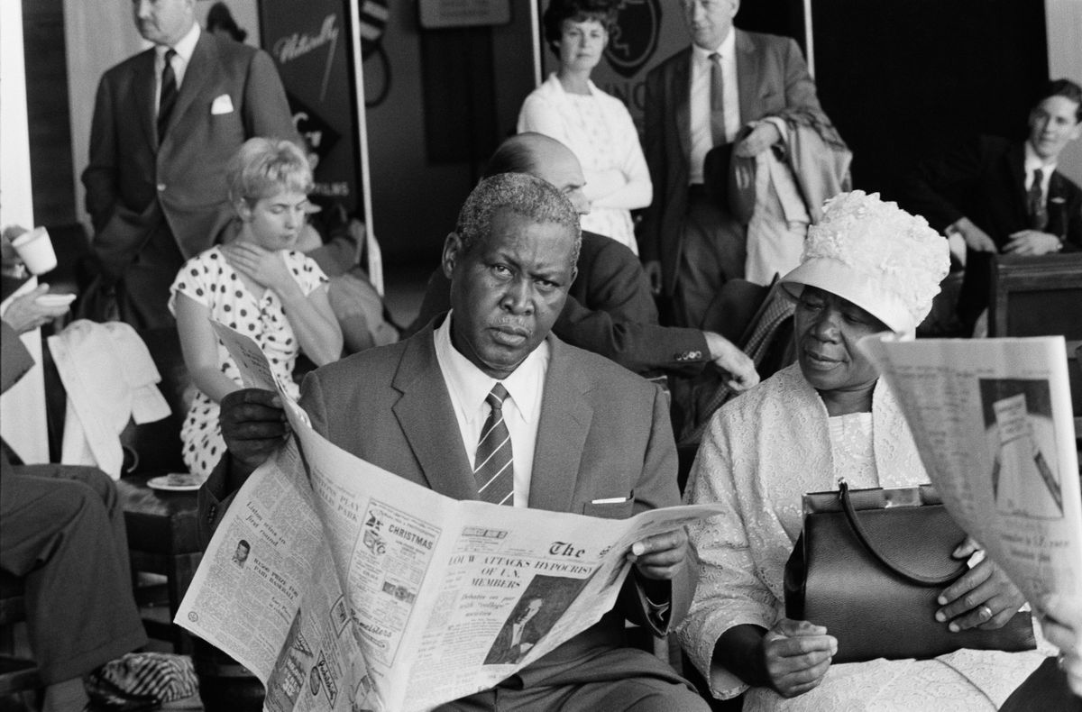 Black and white photograph of Albert Luthuli sat with a newspaper in his hands, as he looks directly into the camera. His wife is sat beside him.