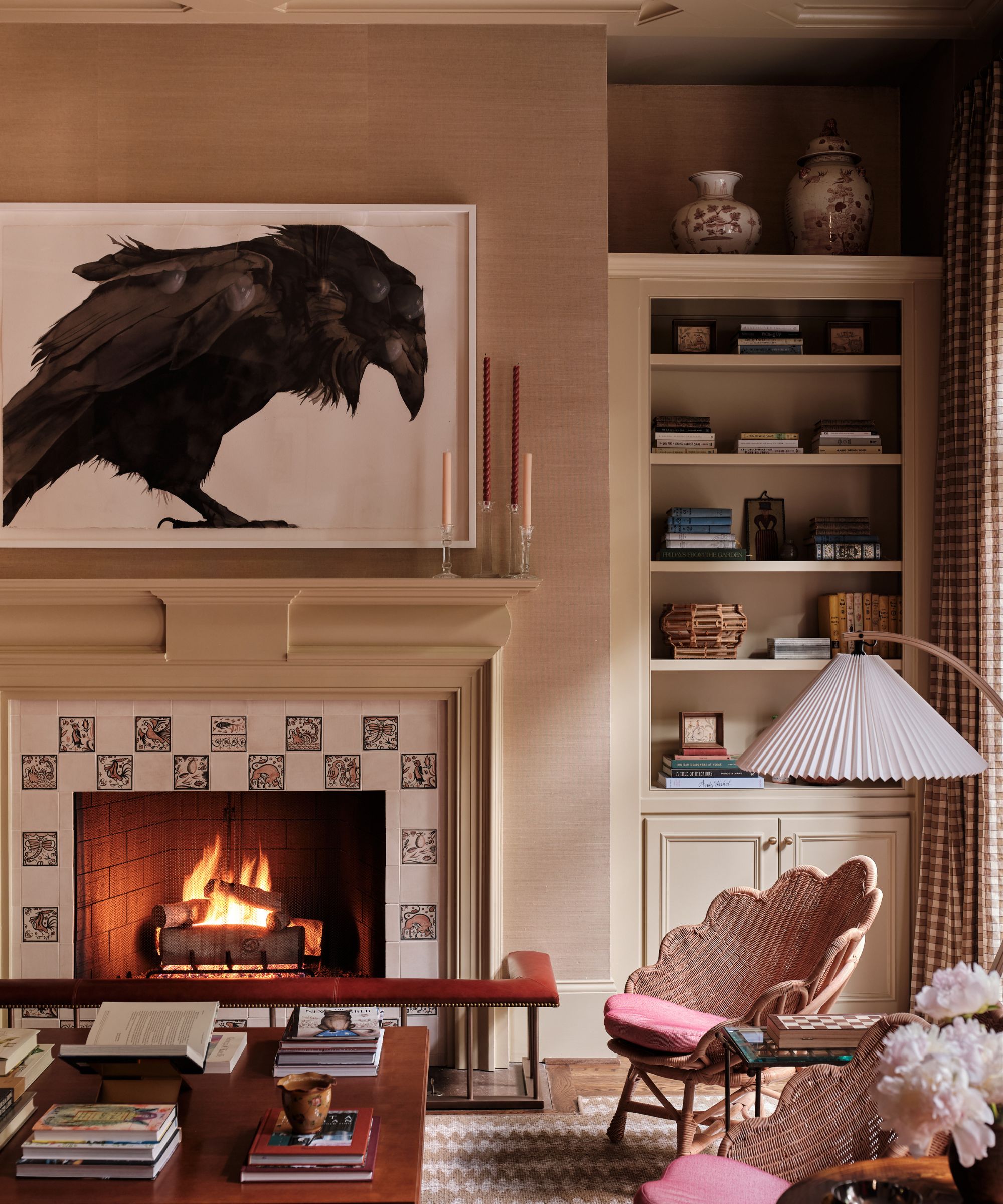 Living room with neutral walls, a tiled fireplace with a red fireplace fender, a built-in bookcase in the alcove, two rattan armchairs, a pleated floor lamp, and a coffee table stacked with books.