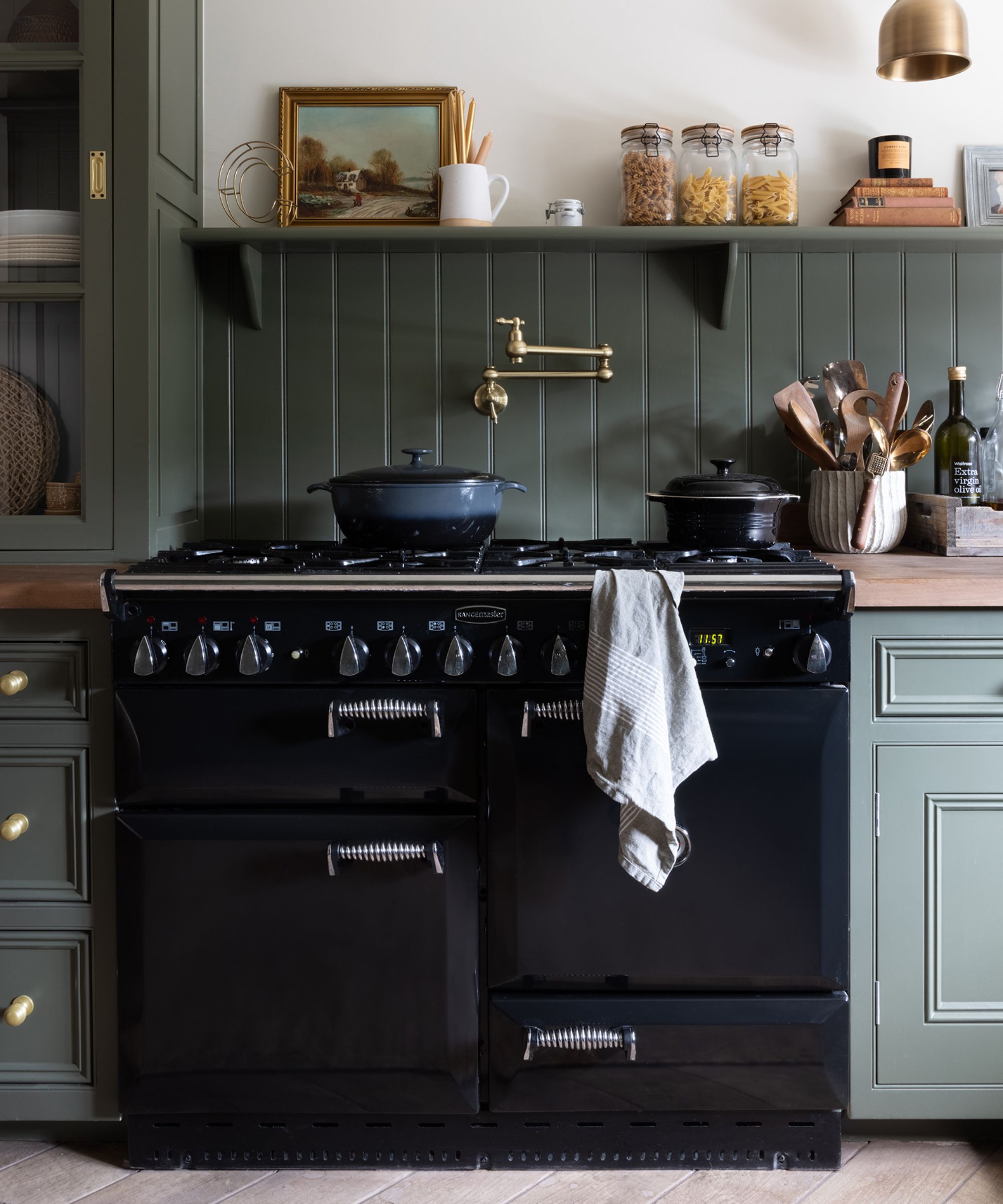 A dark navy range cooker sits between deep sage green cabinets with brass knobs. A pale tea towel hangs from the handle. Above, a matching shelf displays glass jars of pasta, books, and artwork against green panelled walls, lit by warm brass lighting.