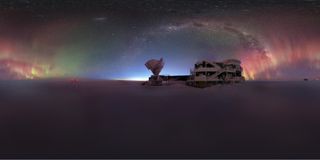 An arch of rainbow colored lights, along with the Milky Way, appear in the night sky above a large rounded building and antenna disk in the snow.
