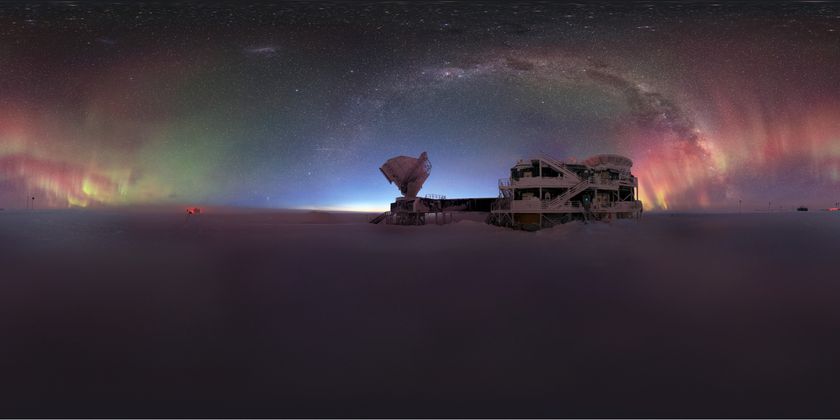 An arch of rainbow colored lights, along with the Milky Way, appear in the night sky above a large rounded building and antenna disk in the snow.
