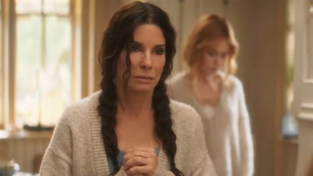 Sandra Bulock in a cardigan and braids clasps her hands and stares directly at the camera. 
