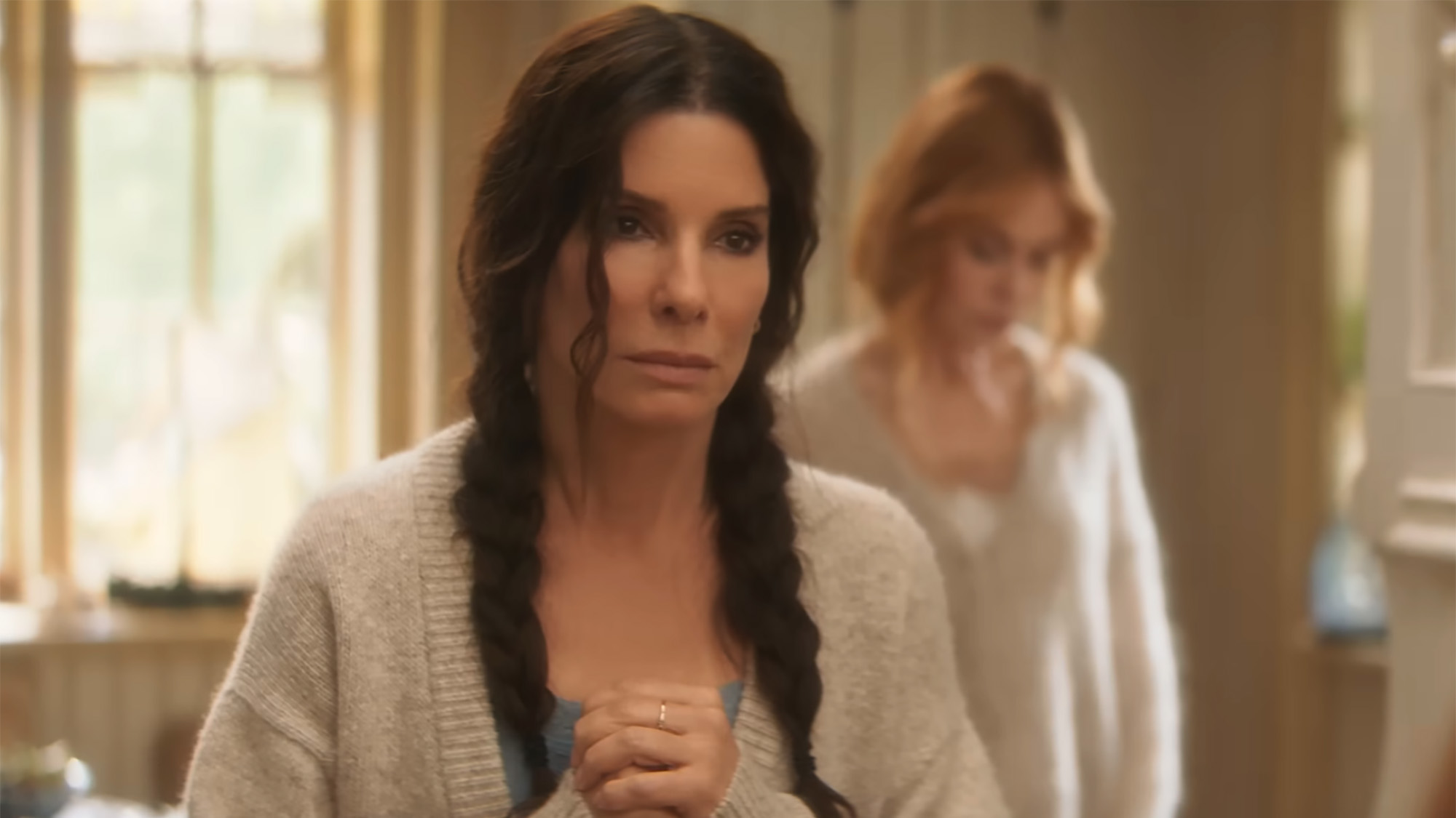 Sandra Bulock in a cardigan and braids clasps her hands and stares directly at the camera. 