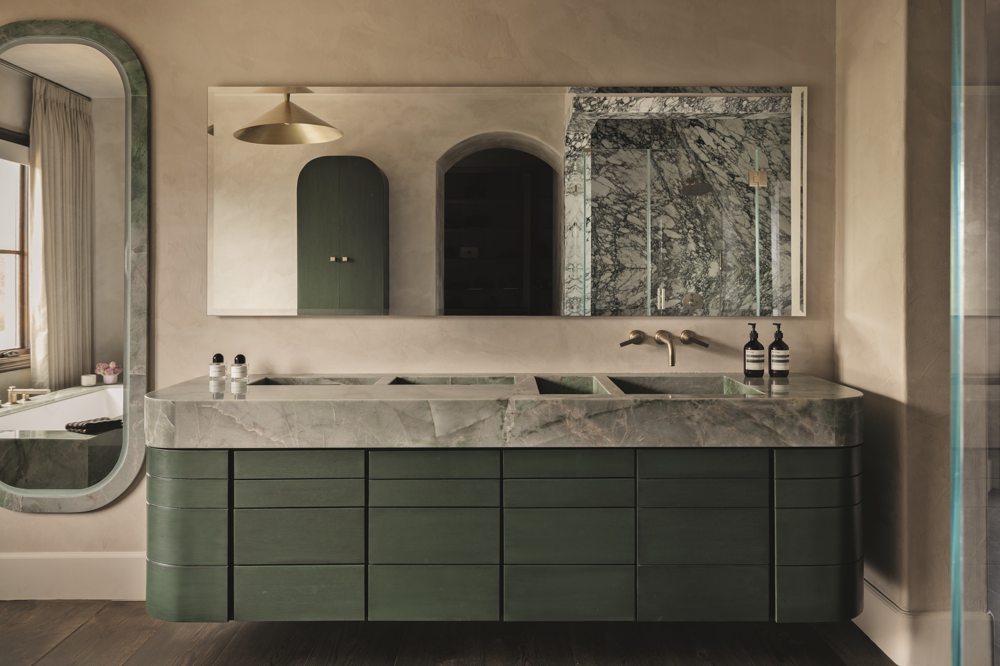 modern bathroom with green floating vanity with gray marble countertop, beige plaster walls, large mirror, large curved floor-length mirror on the wall beside the vanity, dark timber floorboards, and marble shower enclosure