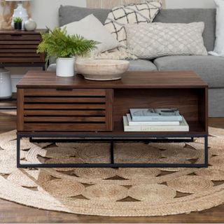 Brown wooden coffee table with slats on one side and a storage shelf on the other, sitting on a black metal frame ontpo of a jute rug in a living room