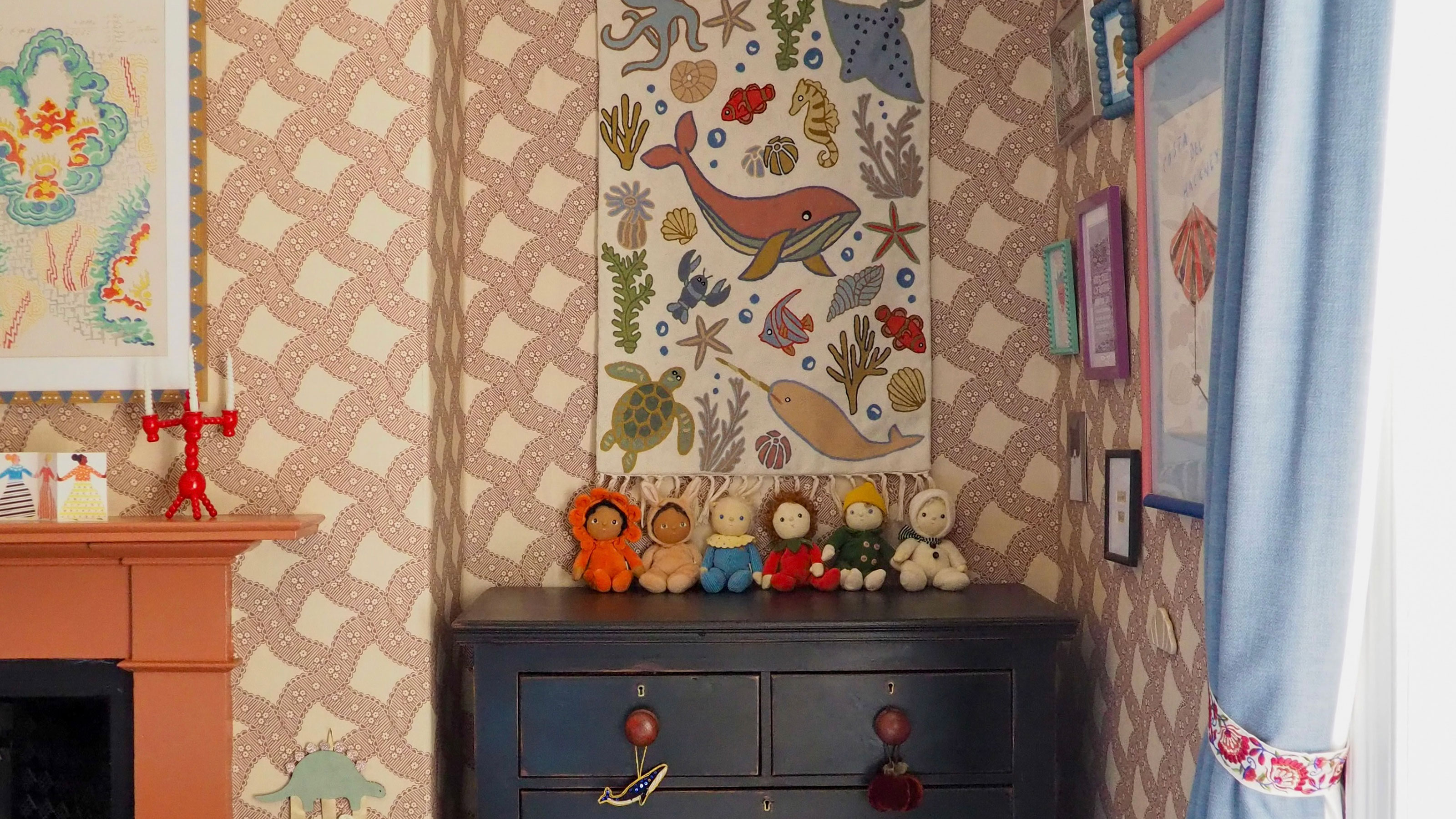 Child's bedroom with patterned wallpaper, dark chest of drawers, blue curtains and colourful wall hangings and prints