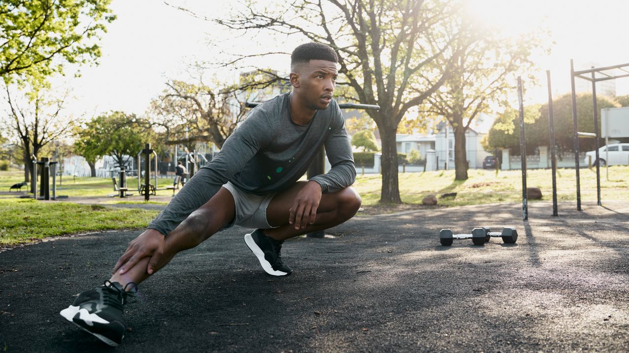 A man in sportswear stretches in a park. He is in a very low squat, with one knee bent and the other leg out to the side. Behind him we see trees and a set of dumbbells.