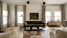 neutral living room with shearling armchairs, a olive green sofa, a round coffee table, timber floors, and three windows with curtains