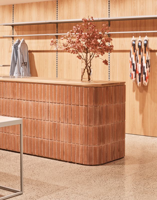 A wood and terracotta tiles-clad fashion boutique boasts a few selected clothing items hung on a minimalist rail, dry flowers, and soft, warm artificial lighting.