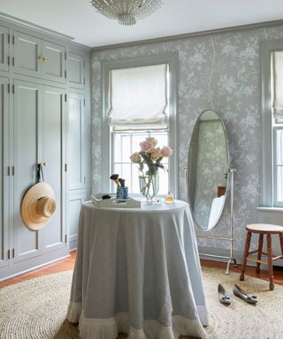 dressing room with skirted table, gray patterned wallpaper and blue gray cabinetry and oval mirror