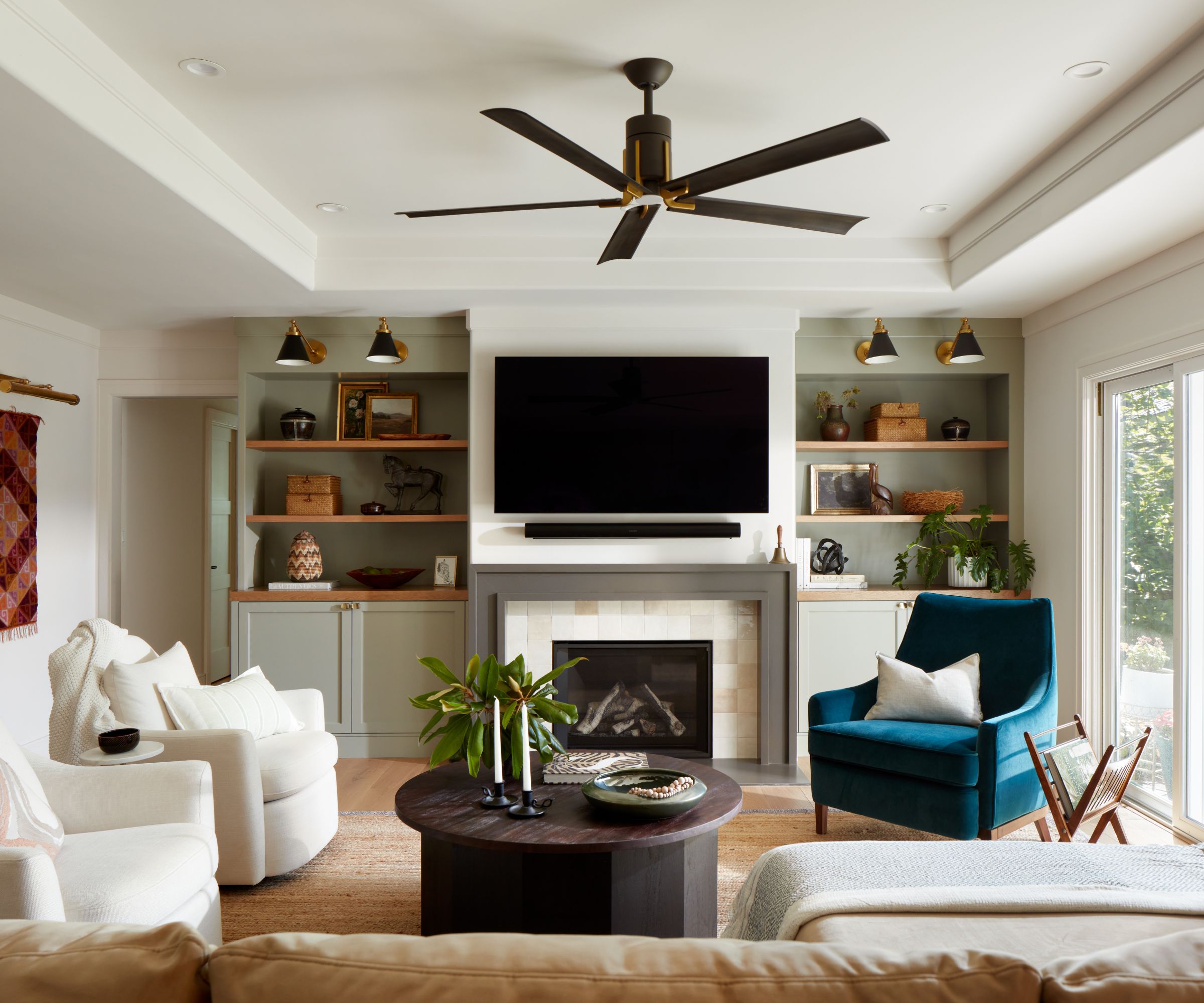 Living room with sage green built-in nooks, a tiled fireplace with a TV above it, a rattan rug with white swivel chairs, a black coffee table, and a blue velvet armchair on top and a ceiling fan above
