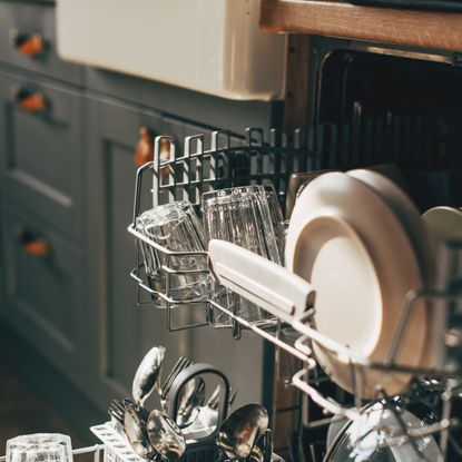 Open dishwasher with loaded top drawer pulled out in a kitchen with grey blue cupboards and a butlers sink