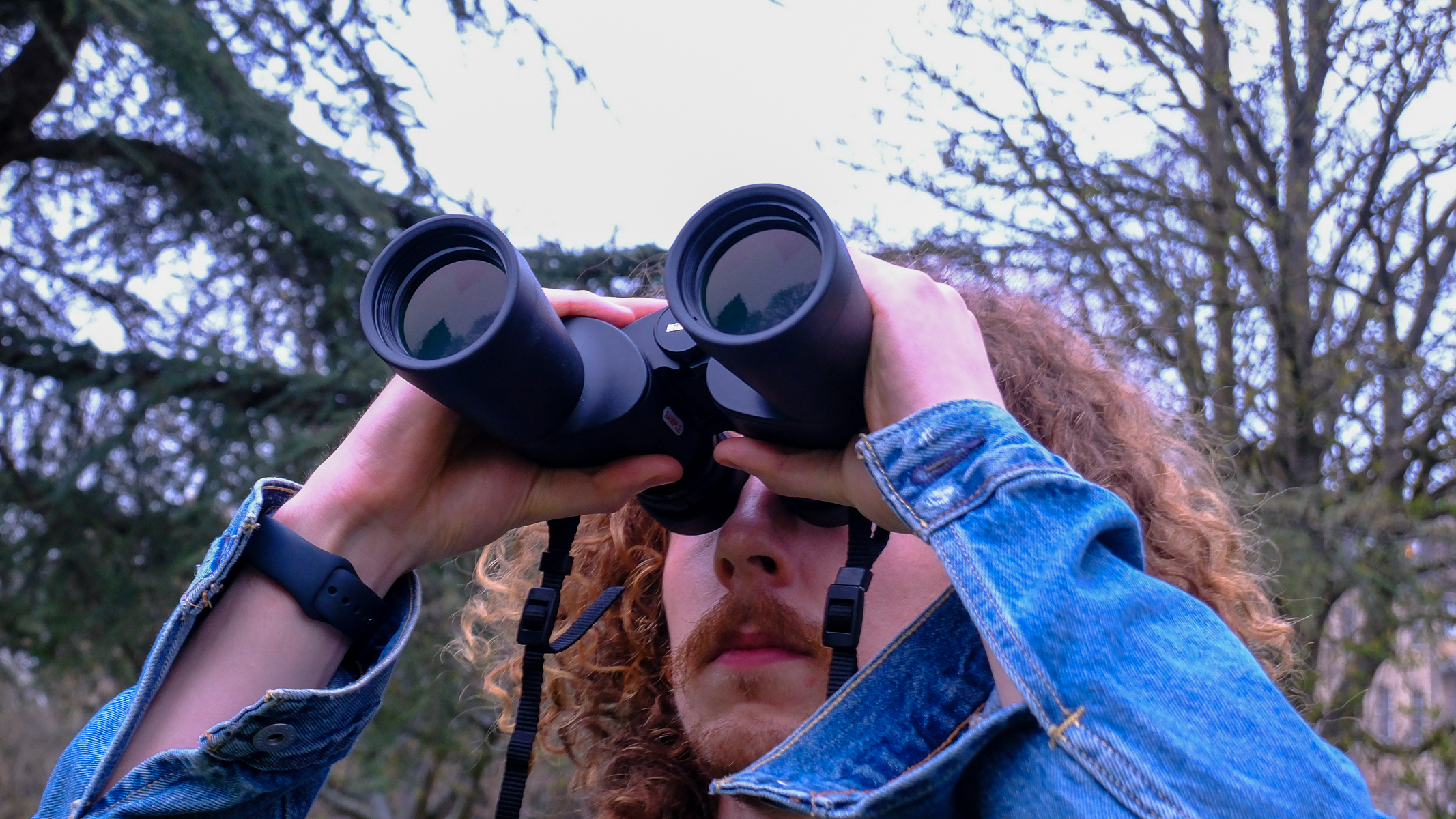 A man looking towards the sky, through the Nikon Action 7x50 binoculars, with trees in the background.