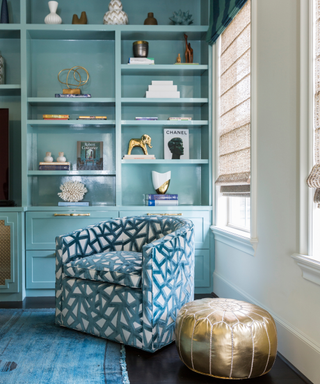 Light blue bookshelf corner with accent chair and gold pouffe next to rug with cobalt tones running through