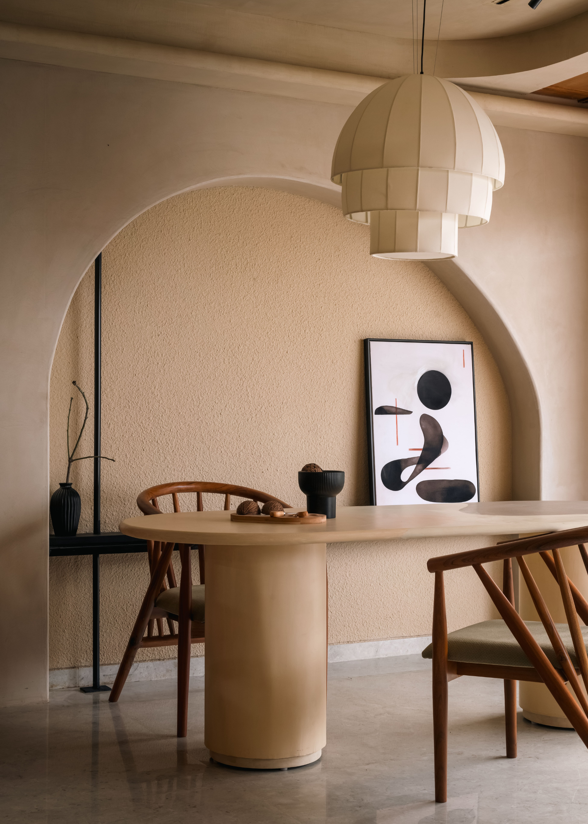 A minimalist dining room with a paper pendant lamp, a beige table, wood armchairs, a pedestal bowl, a cutting board with cut figs beside a leaning painting, and a ceramic vase