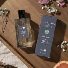 A glass bottle of home fragrance along with a box of home fragrance from ScentAir, on a wooden surface with small sprigs of flowers. 