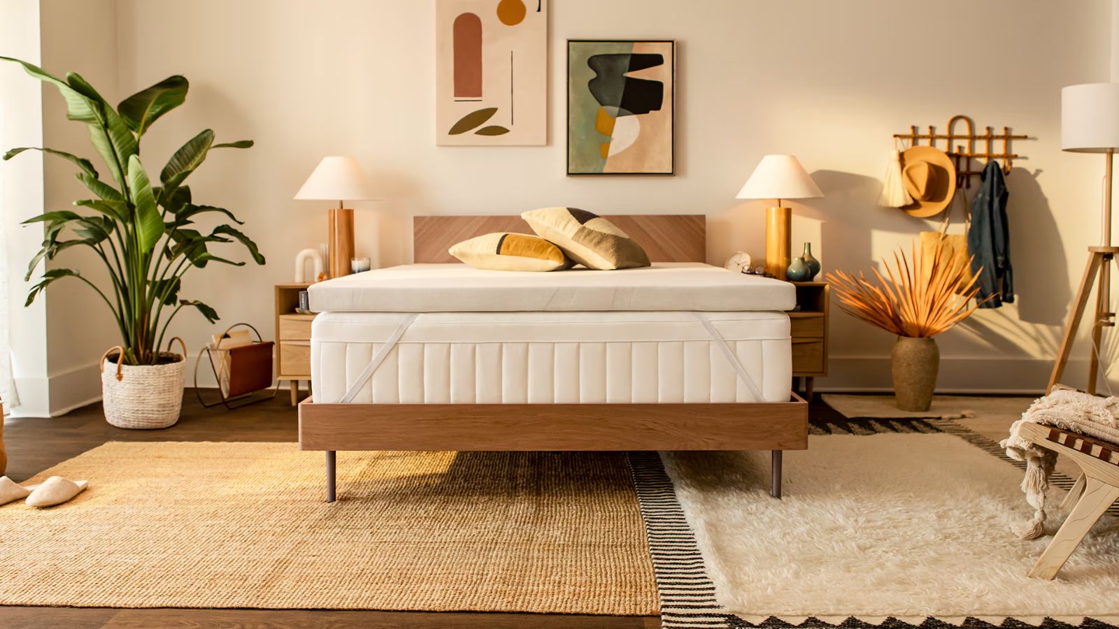 The Tempur-Pedic Adapt Topper shown on a wooden bedframe and white mattress, in a cozy and warm sunlit bedroom space, with mismatched, layered rugs, a large houseplant, abstract artwork above the bed, matching wooden bedside tables and lamps with white shades to either side, and a large coat rack.