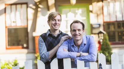 Two smiling gay men standing in the front yard of their home on a sunny day. One man has his arms folded, leaning on the top of a white, picket fence. His partner is beside him.
