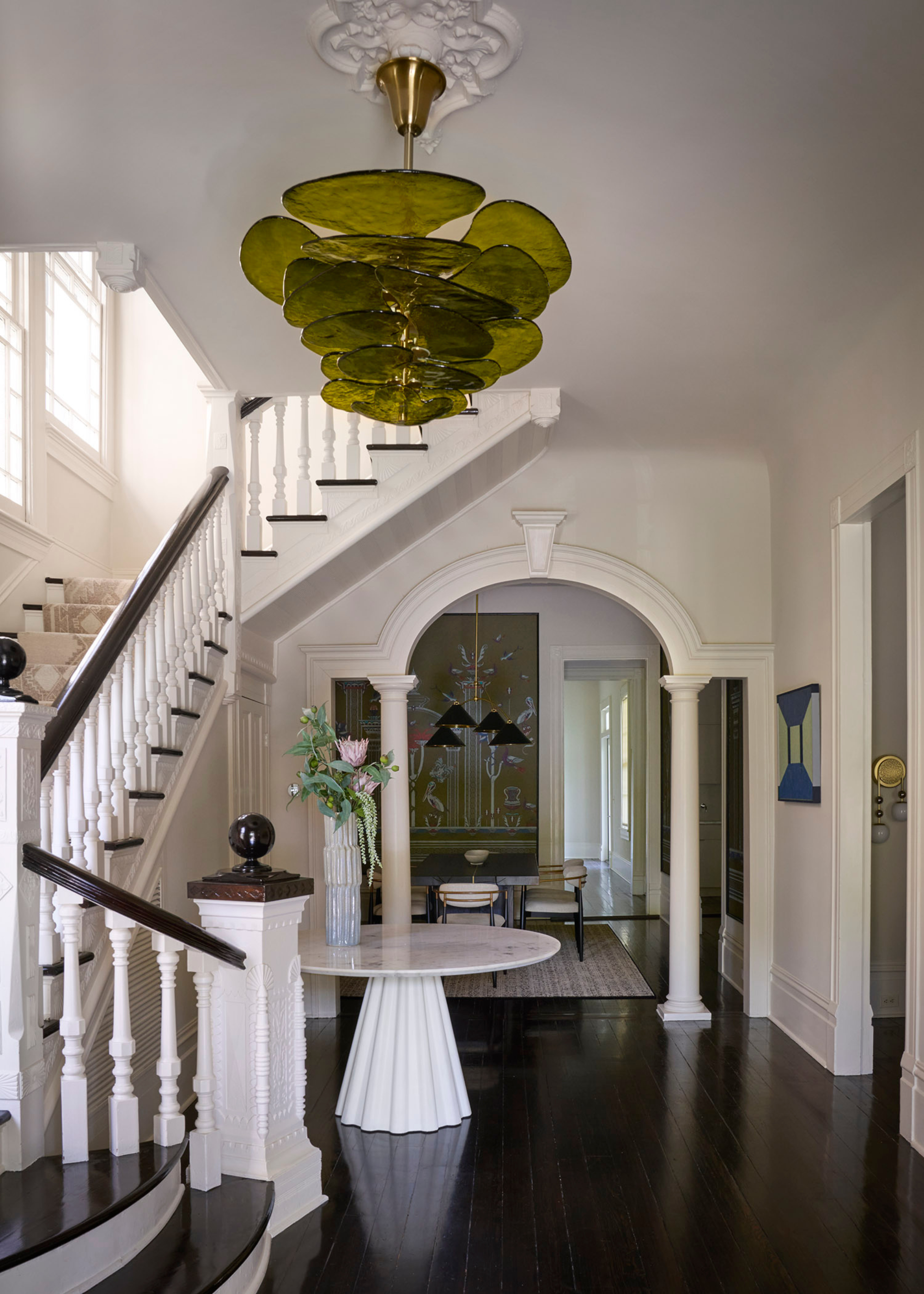 A white entryway with a staircase leading up to the first level. Also pictured are arched doorways, a white side table with a vase of flowers and a green glass chandelier
