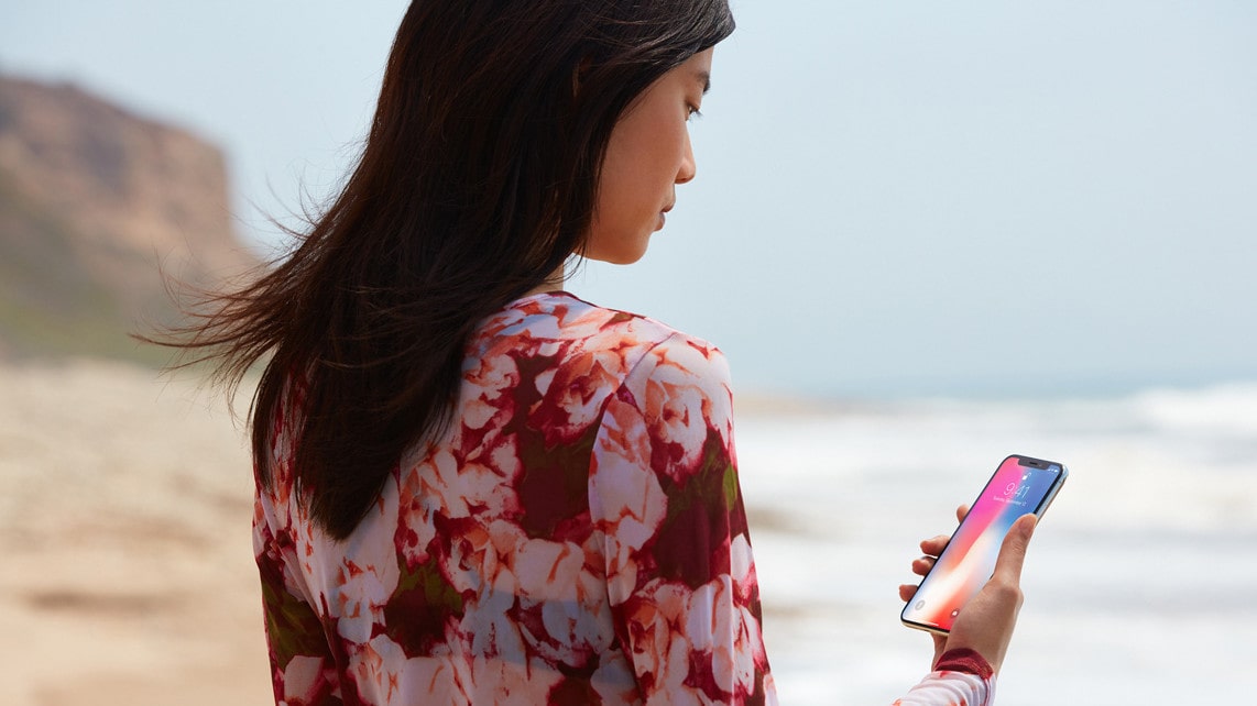 A woman on the beach looks at her iPhone X and unlocks it with Face ID.