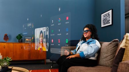 A person wearing smart glasses sits on a couch, surrounded by holographic menus and screens projected in front of them.