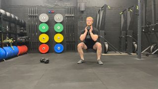 Fitness writer Harry Bullmore performs a goblet squat with a dumbbell clasped to his chest. He's in a gym, with colorful weight plates stacked behind him.