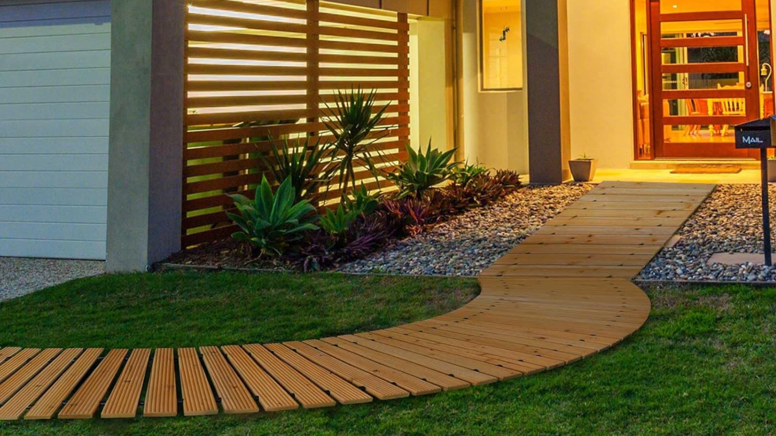 A curved wooden garden pathway on grass leads to a straight wooden pathway on stone that leads to a home's red glass-framed front door, lit up in the evening, with a garage with fence panels on one side, with plants running along it