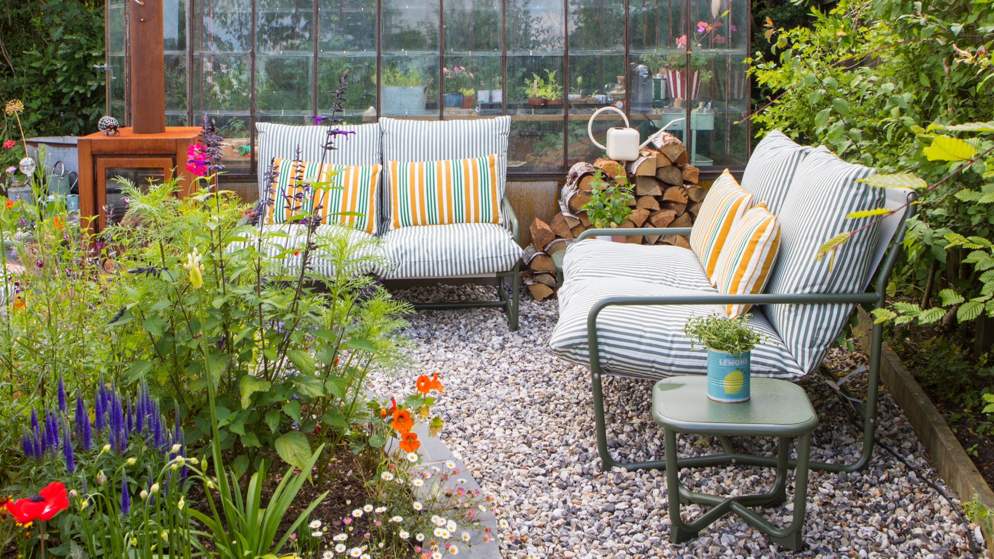 A gravel garden with two John Lewis Marcy 2-seater sofas in green stripe in front of a greenhouse