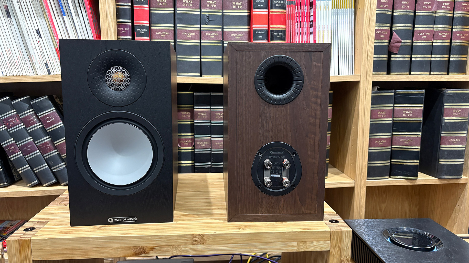 Monitor Audio Bronze 50 7G standmount speakers on wooden hi-fi rack in front of bookcase, one speaker is rear-facing showing binding posts