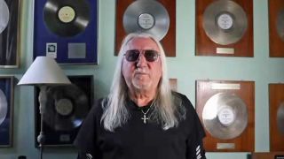 Mick Box speaking in front of a wall of gold discs