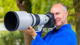 A person holding a large Canon super telephoto camera with a long lens, set against a backdrop of lush greenery