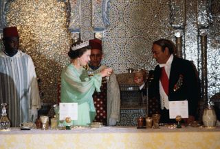 Queen Elizabeth wearing a green gown and diamond tiara raising a glass to King Hassan of Morocco at a banquet table