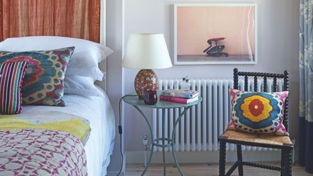 A colorful bedroom with a white bed with colorful cushions and blankets, a black chair, a clear circular table with a colorful lamp with white shade, and a large artwork hung above the radiator.