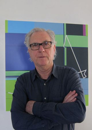 Headshot photograph of Manfred Mohr, wearing a dark blue shirt, arms folded, light grey hair and black framed glasses, stood in front of a colourful piece of computerised artwork on a white wall