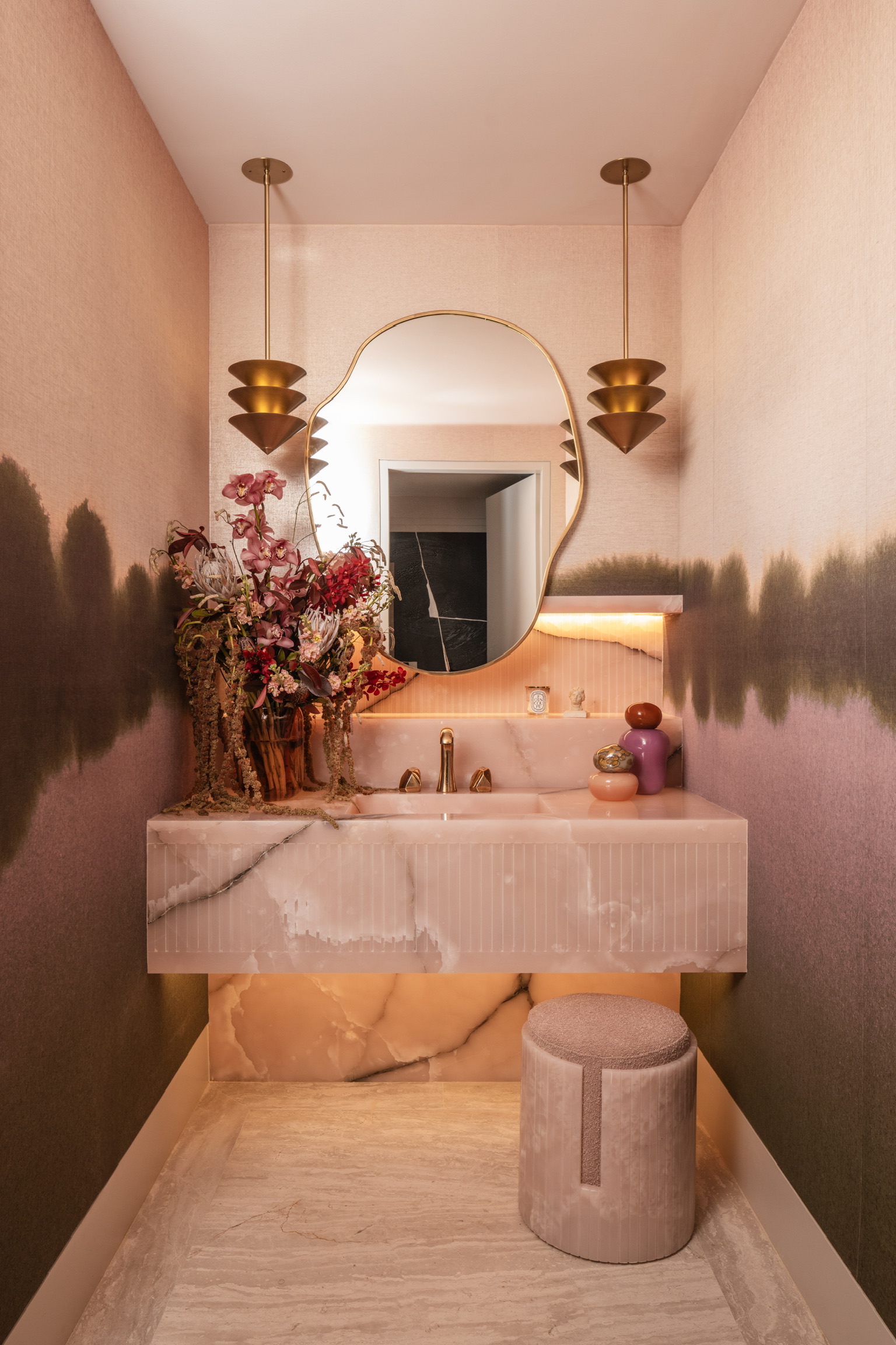 a light pink bathroom with a wall mural of a lake, a pink onyx vanity, gold mirror and lights, all lit by hidden lighting with a warm glow